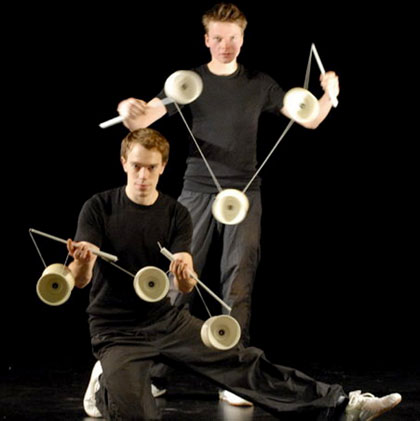 Un artiste accroupi et l'autre debout jonglent avec plusieurs diabolos a la fois face au public
