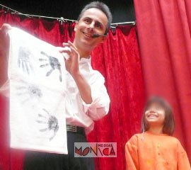 Cet artiste magicien avec son micro presente un tissu blans avec des empreintes de mains devant un enfant ebahi