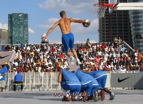 Un basketteur fait un smash lors d’un spectacle de dunk basket en prenant appui sur trois basketteurs accroupis sous le panier